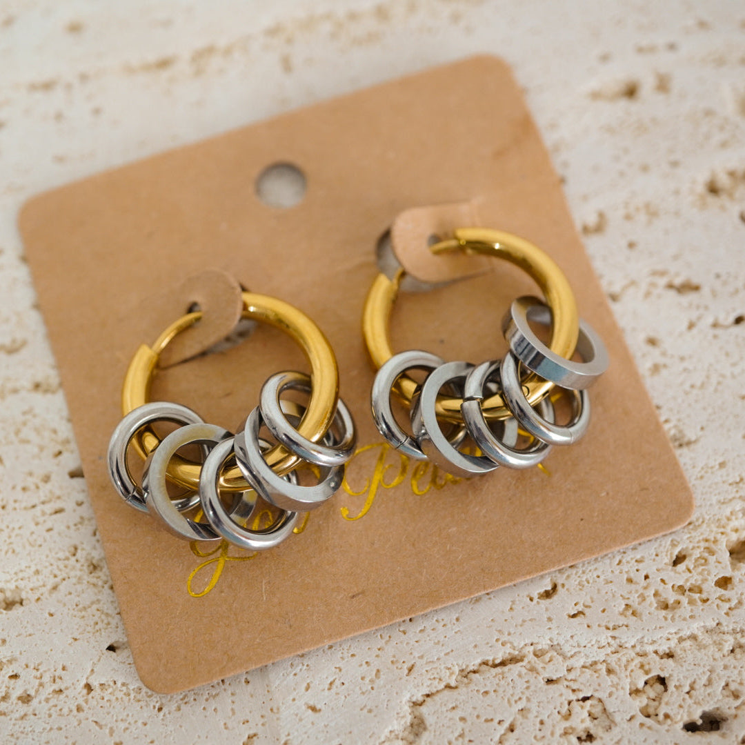 A pair of gold and silver hoop earrings with layered interlocking rings, displayed on a brown kraft paper card over a textured beige stone surface.