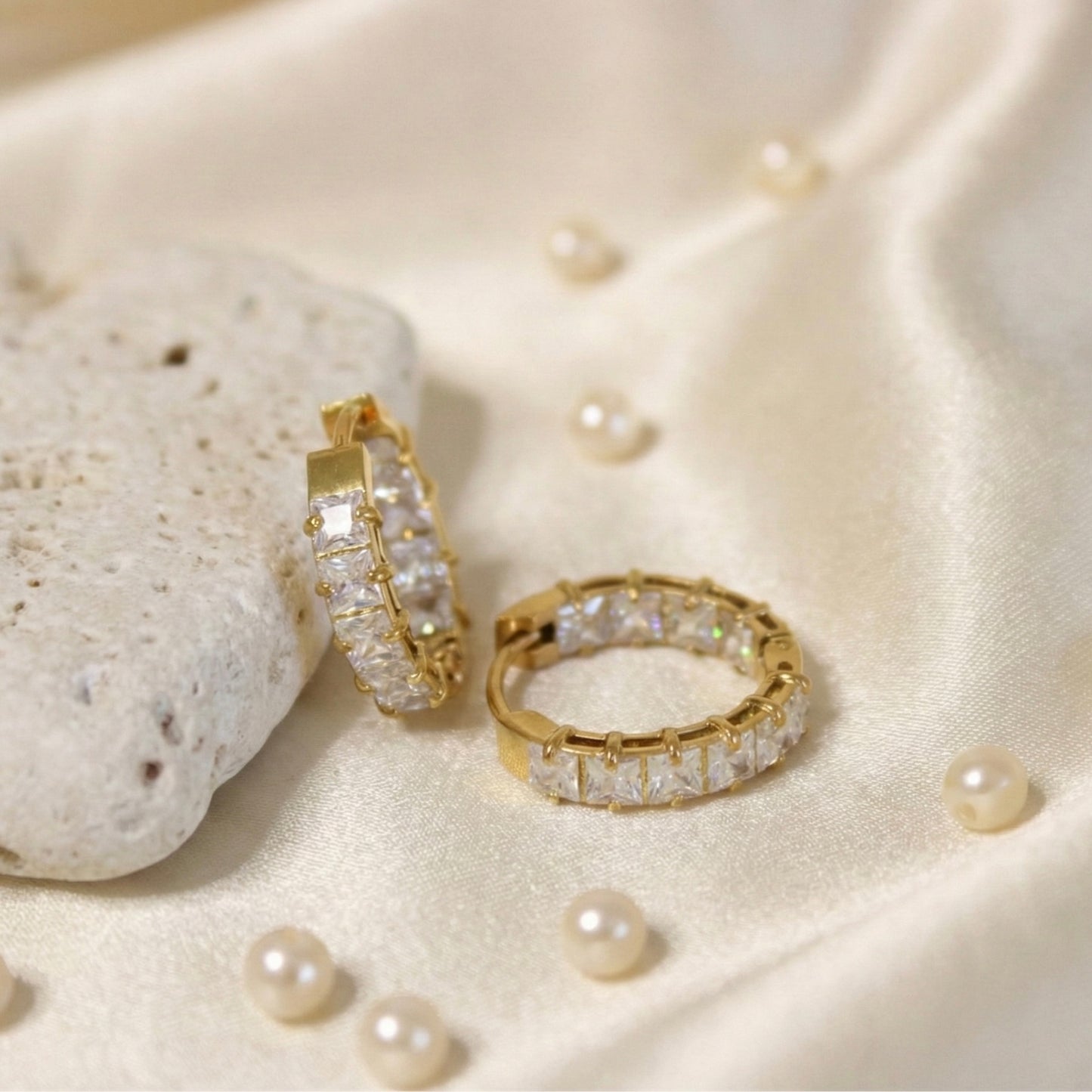 Luxurious overhead lifestyle photography of gold square-set huggie earrings resting on draped off-white satin fabric scattered with small pearls and a white stone.