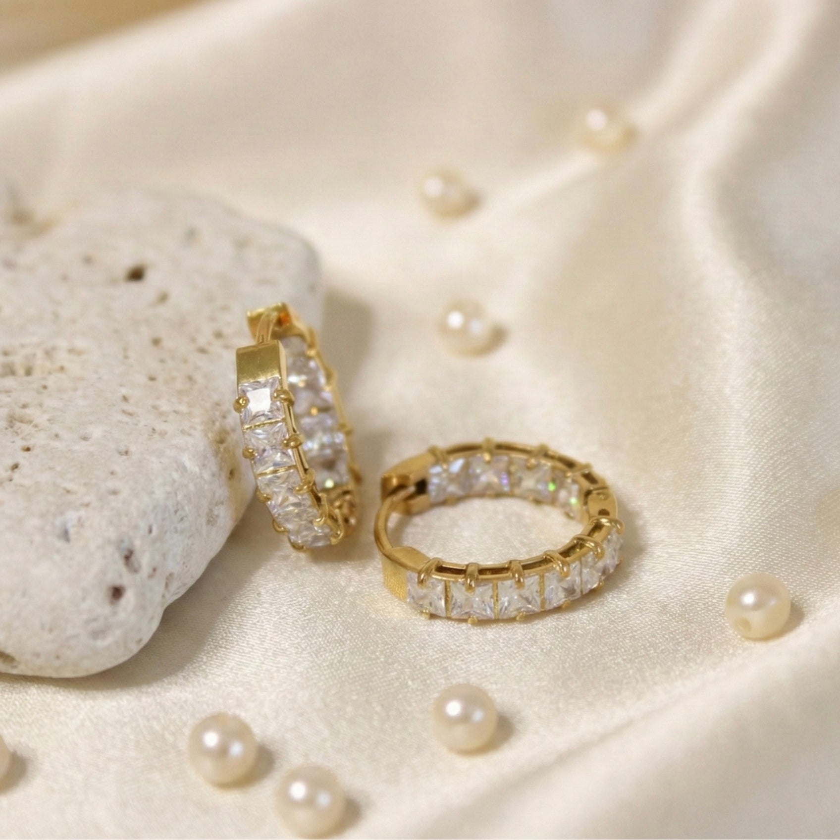 Luxurious overhead lifestyle photography of gold square-set huggie earrings resting on draped off-white satin fabric scattered with small pearls and a white stone.