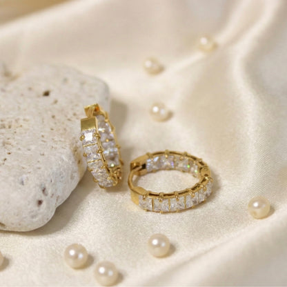 Luxurious overhead lifestyle photography of gold square-set huggie earrings resting on draped off-white satin fabric scattered with small pearls and a white stone.