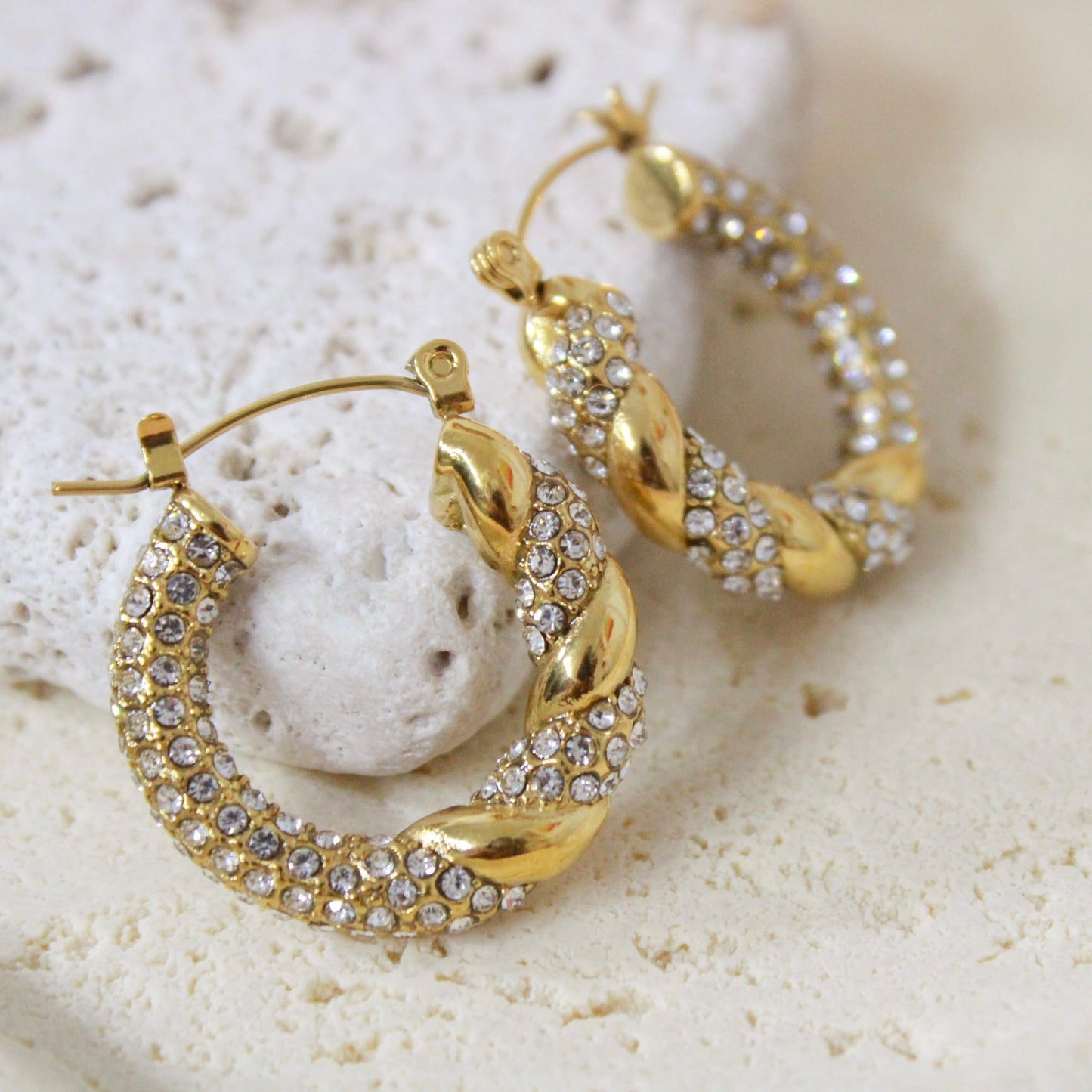 Close up of gold twisted huggie hoop earrings with small pave crystals resting on porous white stone