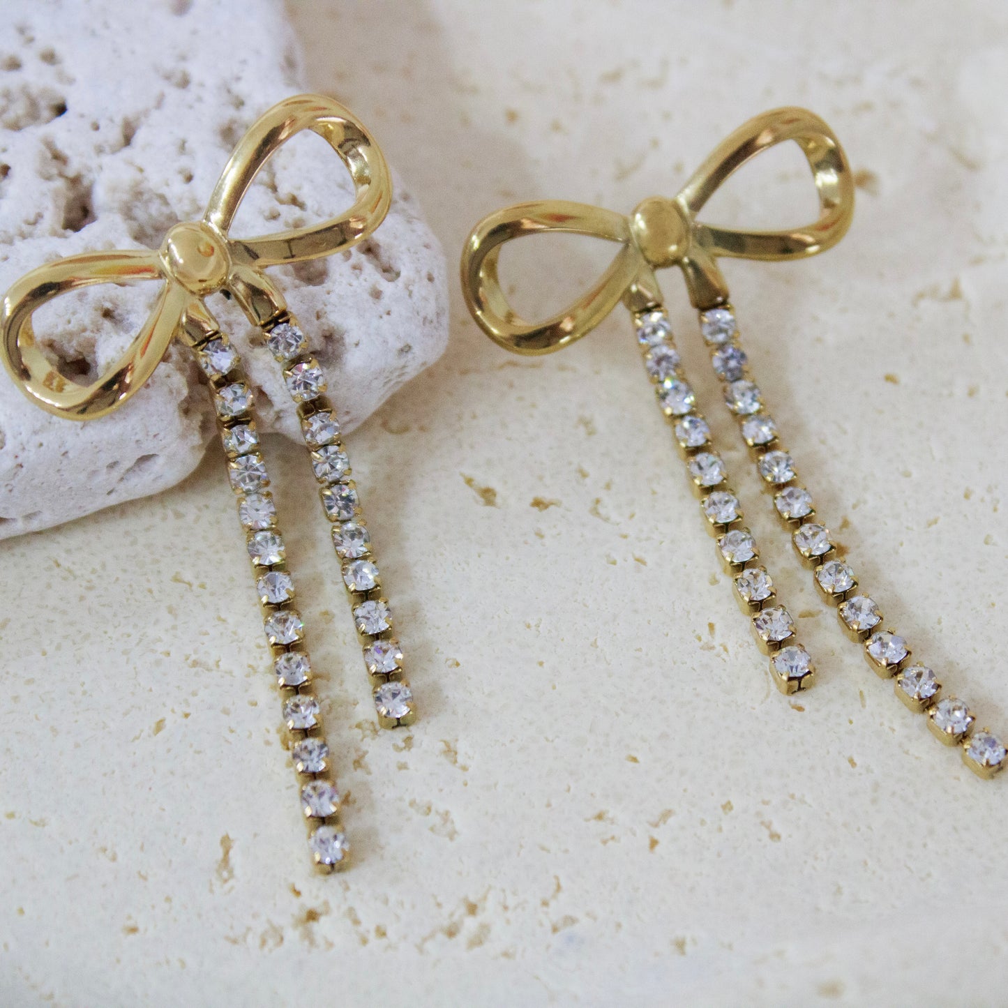 Close-up of 18K gold plated ribbon bow earrings with sparkling cubic zirconia fringe resting on a textured white stone surface. Elegant waterproof jewelry