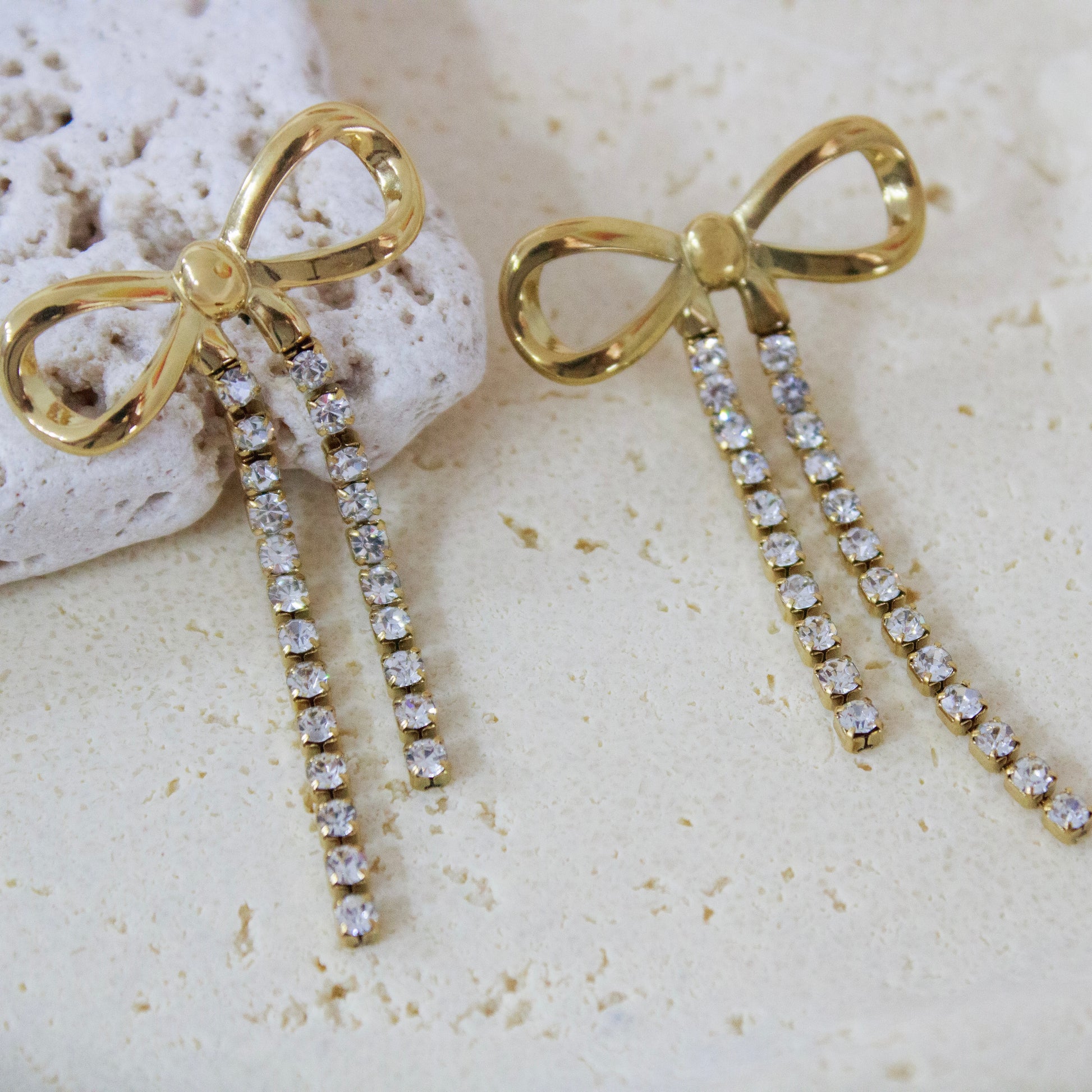 Close-up of 18K gold plated ribbon bow earrings with sparkling cubic zirconia fringe resting on a textured white stone surface. Elegant waterproof jewelry