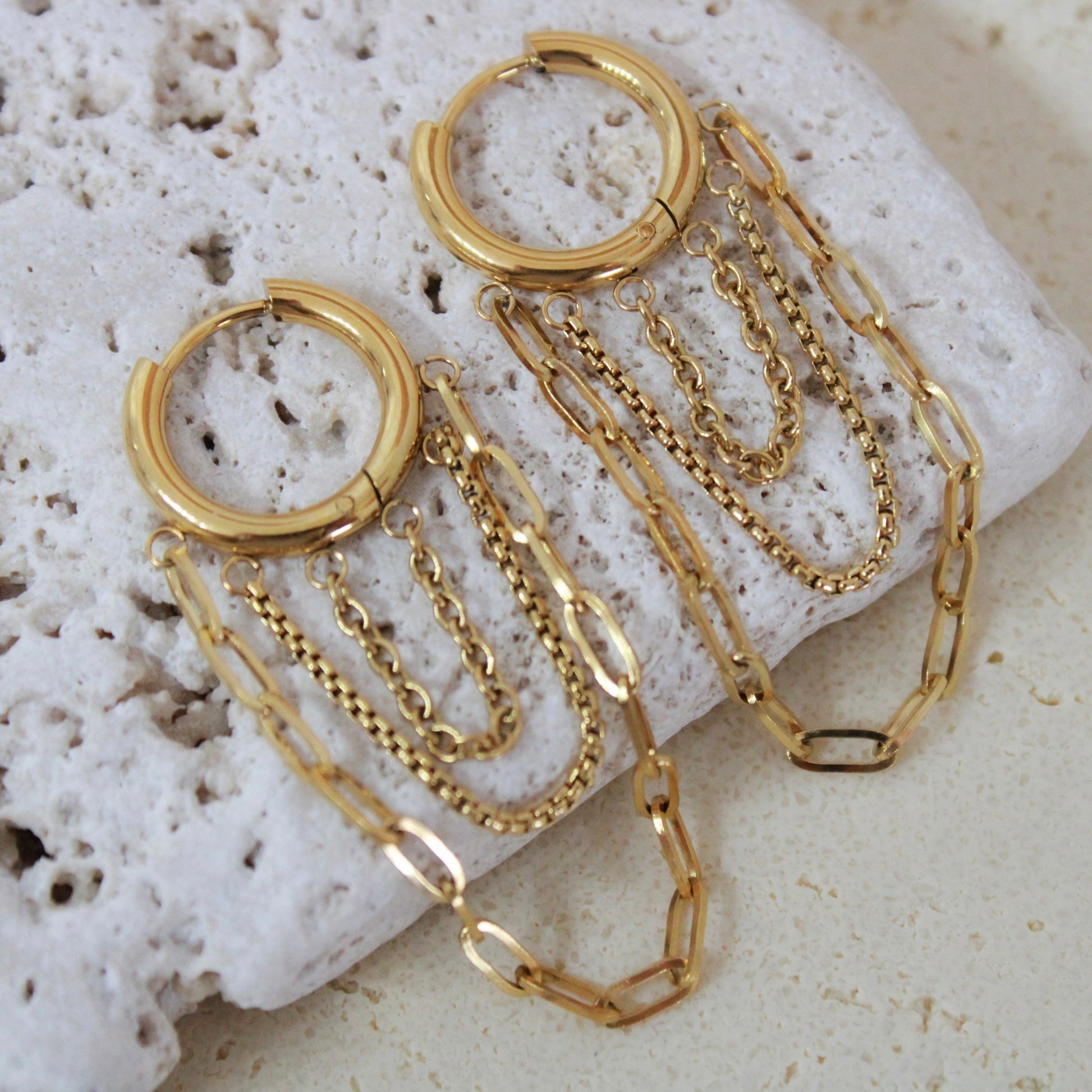 Luxury gold waterproof earrings featuring layered fringe chains. Hypoallergenic stainless steel jewelry on a textured stone background.
