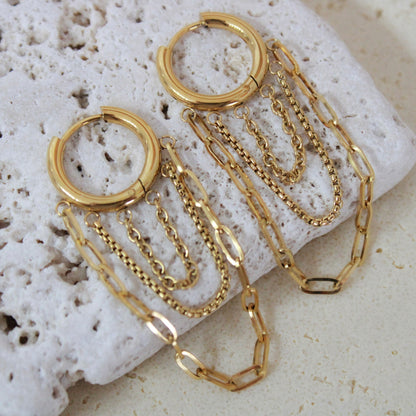 Luxury gold waterproof earrings featuring layered fringe chains. Hypoallergenic stainless steel jewelry on a textured stone background.
