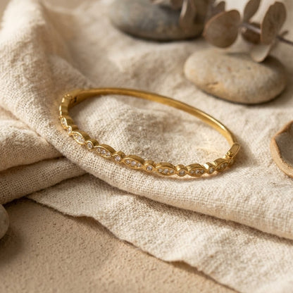 A gold bangle bracelet with a row of small clear stones, displayed on a textured beige linen cloth with decorative stones and dried leaves.