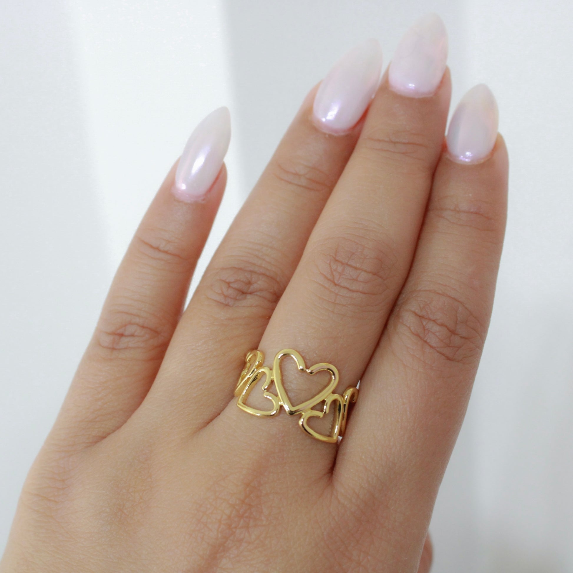 Gold heart-shaped ring on a hand with pink nail polish against a white background